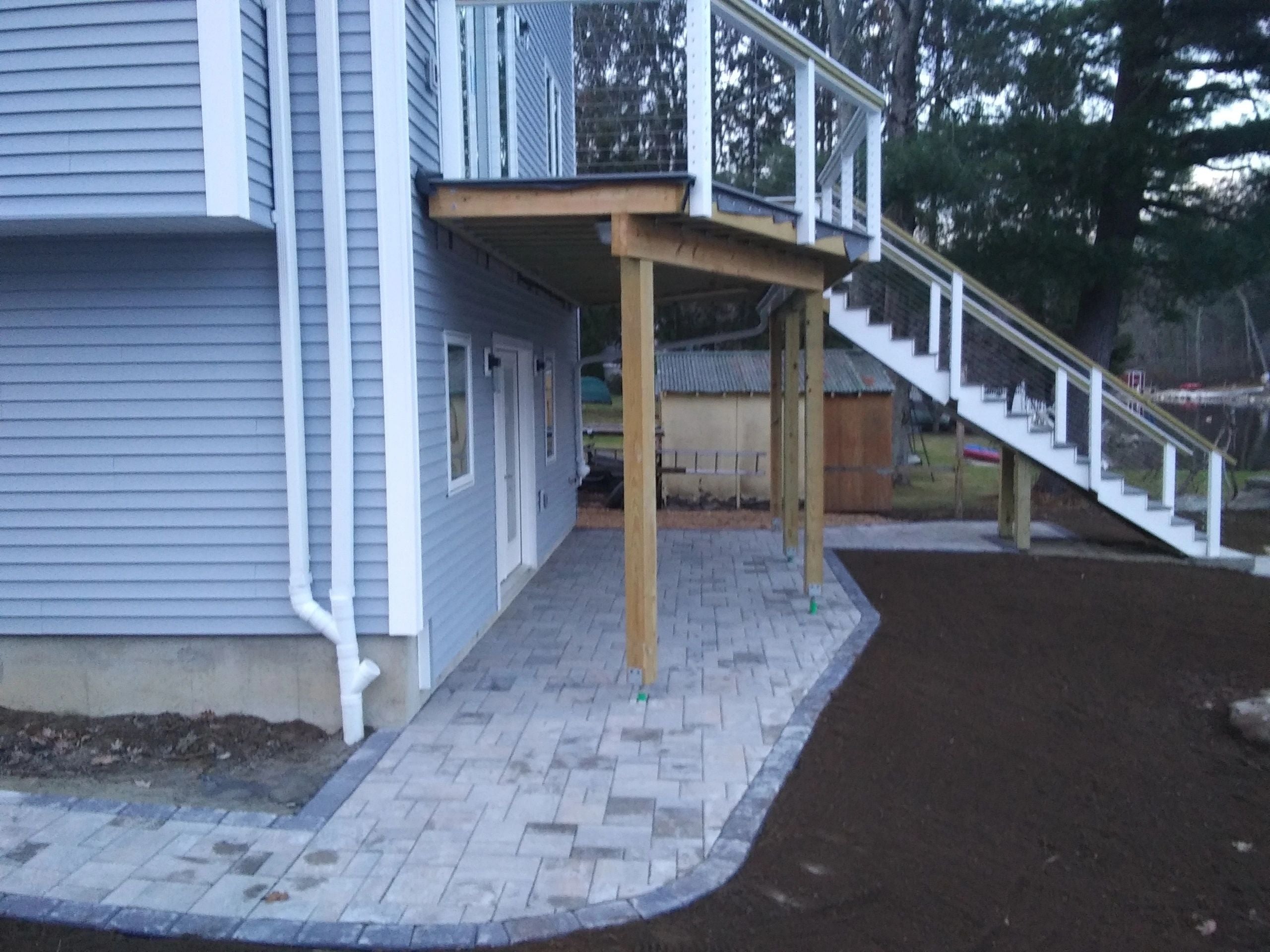 Paver patio tucked under a raised deck at a Charlton lake house, surrounded by fresh loam and landscaping. Project by KRD Landscaping.