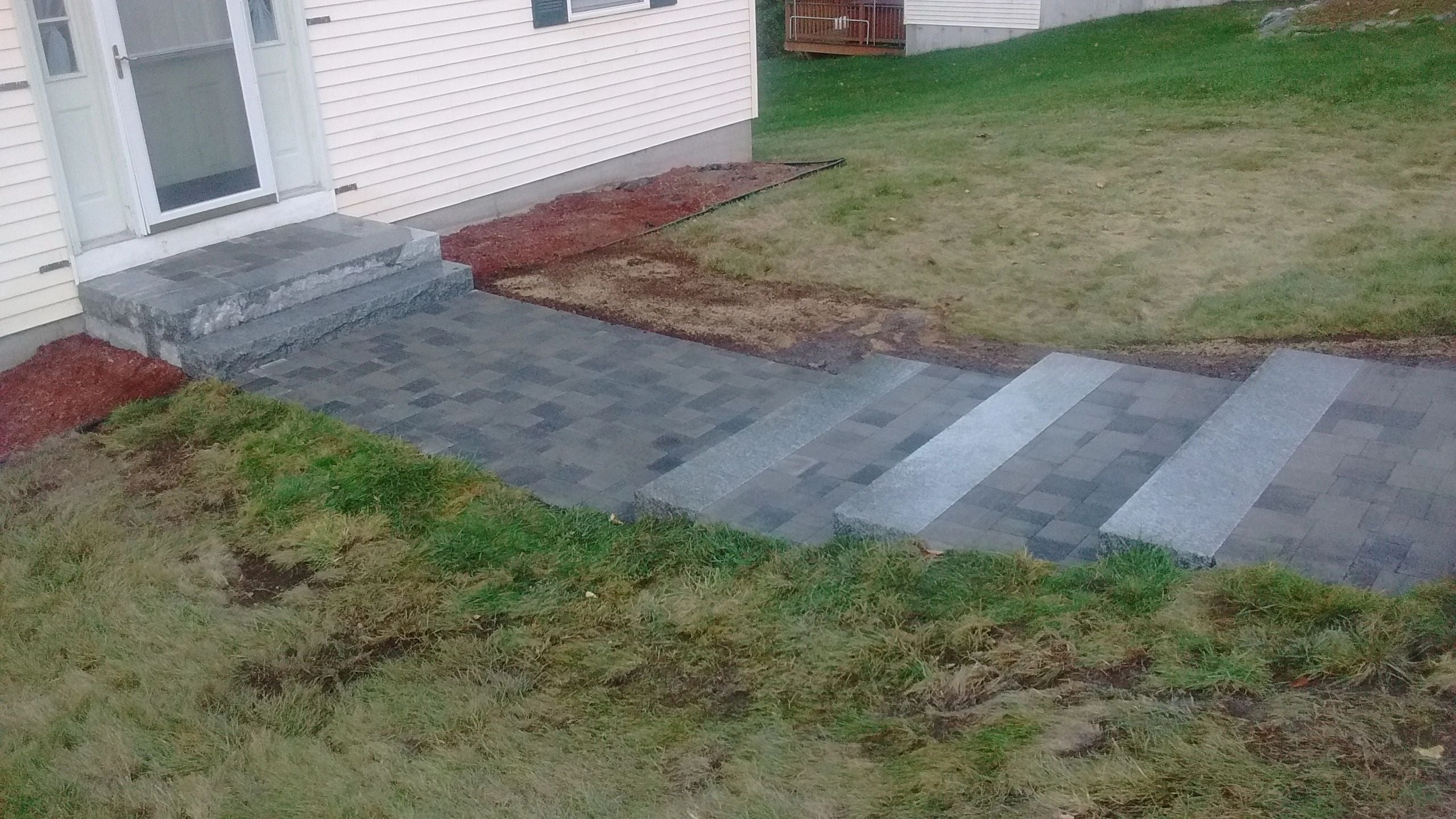 Completed entryway in Webster, MA, featuring granite steps, a paver landing, and a walkway leading down a grassy slope, surrounded by fresh mulch. Project by KRD Landscaping.
