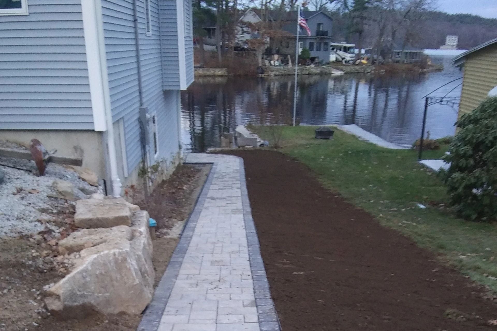Newly installed paver walkway running along the waterfront of a lake house in Charlton, bordered by fresh loam and overlooking the water. Project by KRD Landscaping.
