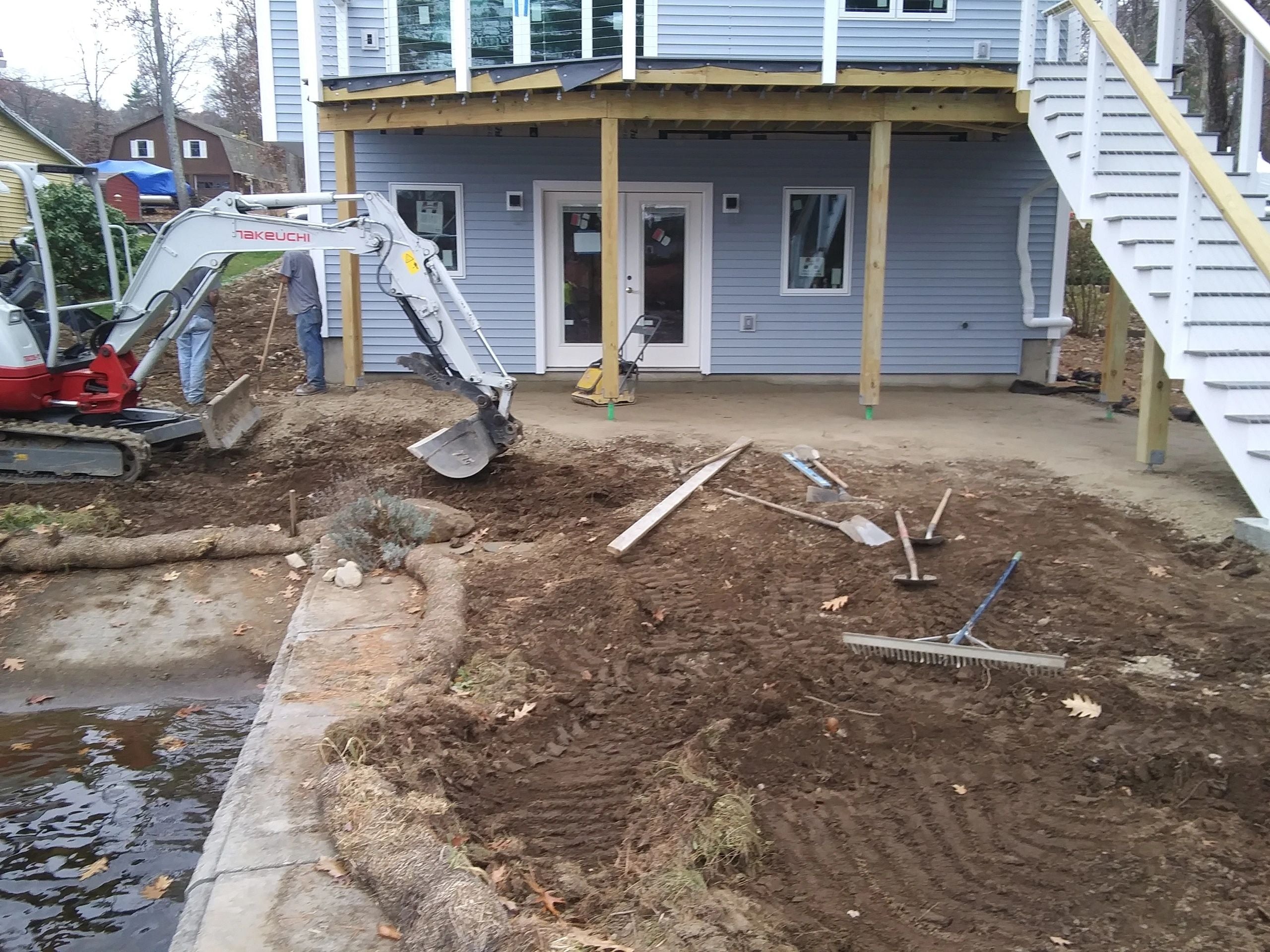 Grading work in progress at a lake house in Charlton, featuring an excavator, disturbed soil, and workers preparing the ground for a paver walkway and patio. Project by KRD Landscaping.