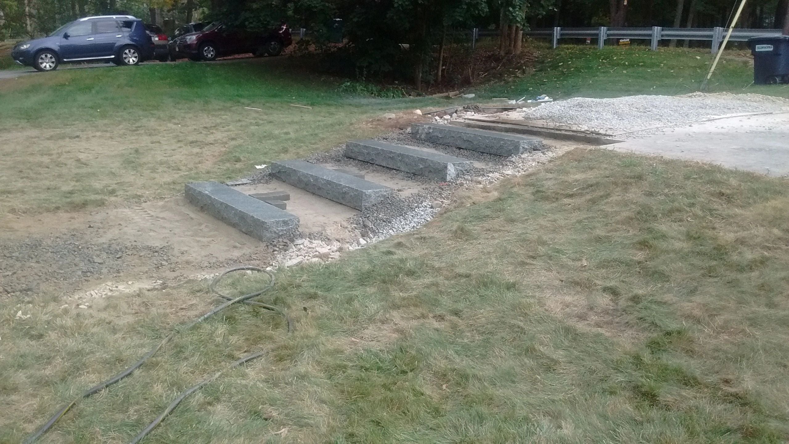 Granite steps under construction on a grassy slope, surrounded by loose gravel, dirt, and construction materials, with a guardrail, parked cars, and trees in the background.