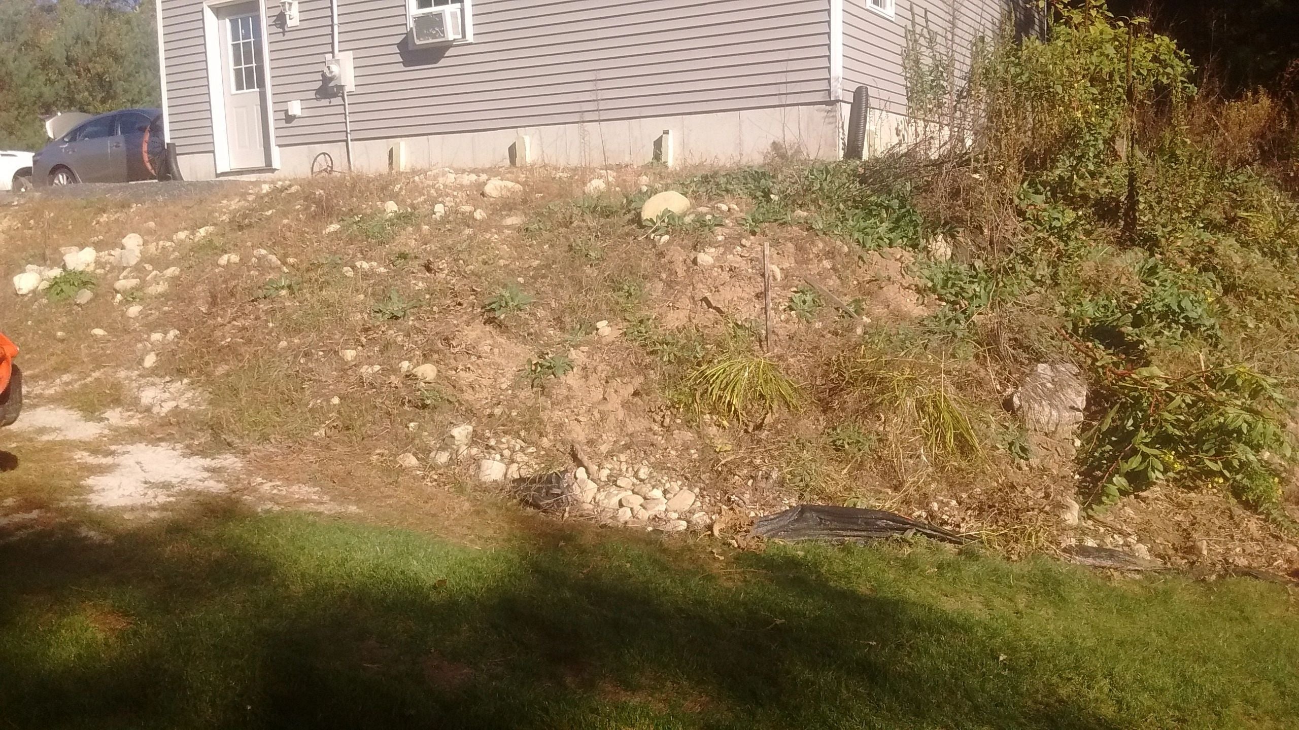 A residential property in Dudley before landscaping, showing a steep, rocky slope with dry grass and scattered stones leading up to a house with light gray siding. The terrain is unlandscaped and uneven.
