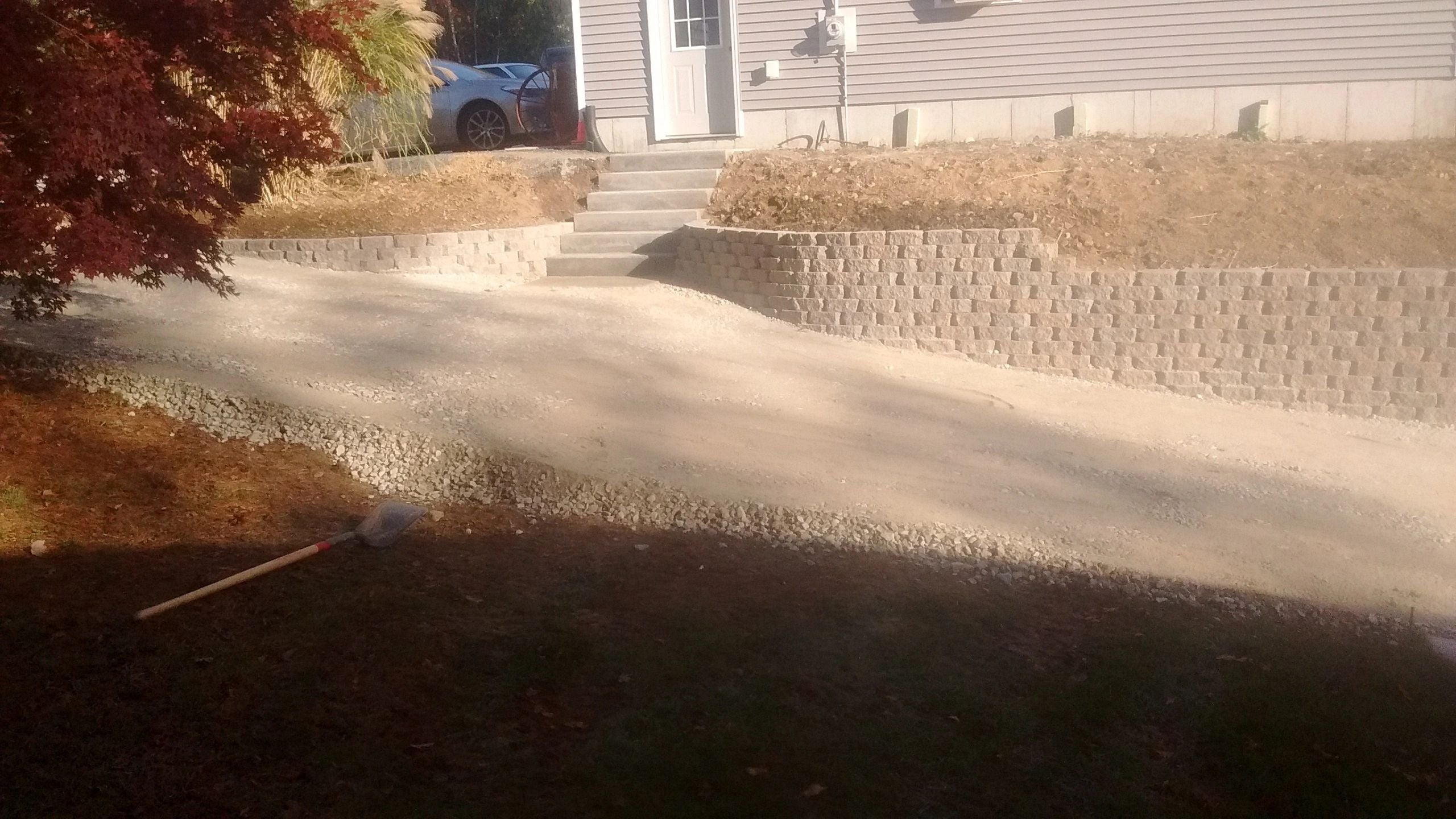 A completed residential landscaping project in Dudley featuring a curved tan masonry retaining wall, a gravel driveway, and concrete stairs leading to a house with light-colored siding. A Japanese Maple tree with red leaves adds vibrant color to the scene.