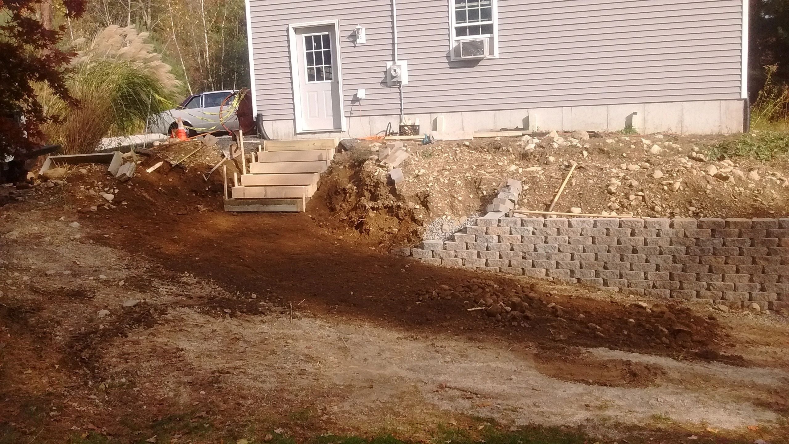 A residential property in Dudley showcasing a partially constructed retaining wall made of gray concrete blocks, adjacent to a wooden staircase leading to a house with light gray siding. The yard is under construction with visible dirt and gravel.