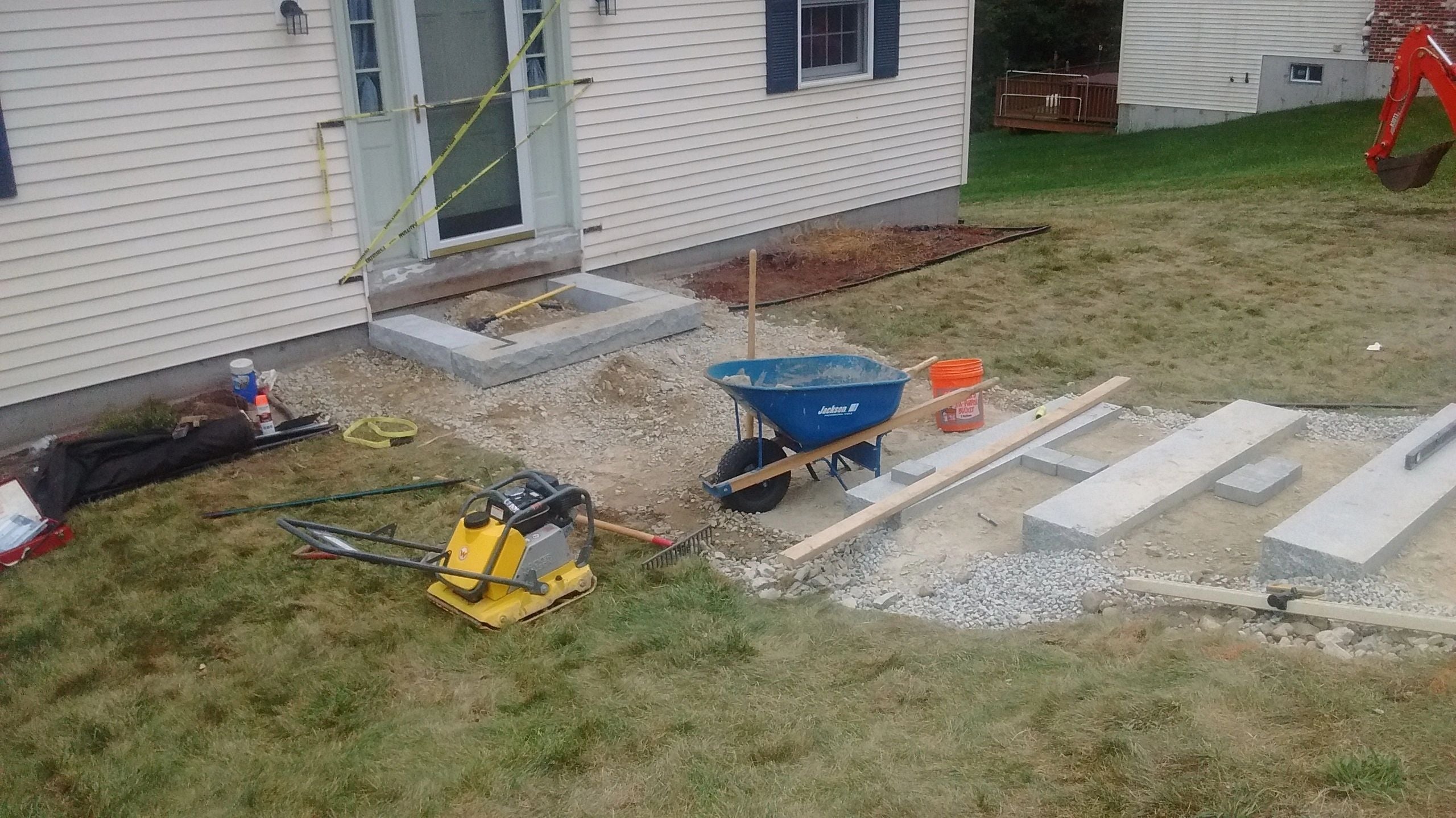 Construction site in Webster, MA, showing granite foundation blocks, gravel, and tools like a wheelbarrow and plate compactor, with caution tape marking the area. Project by KRD Landscaping.