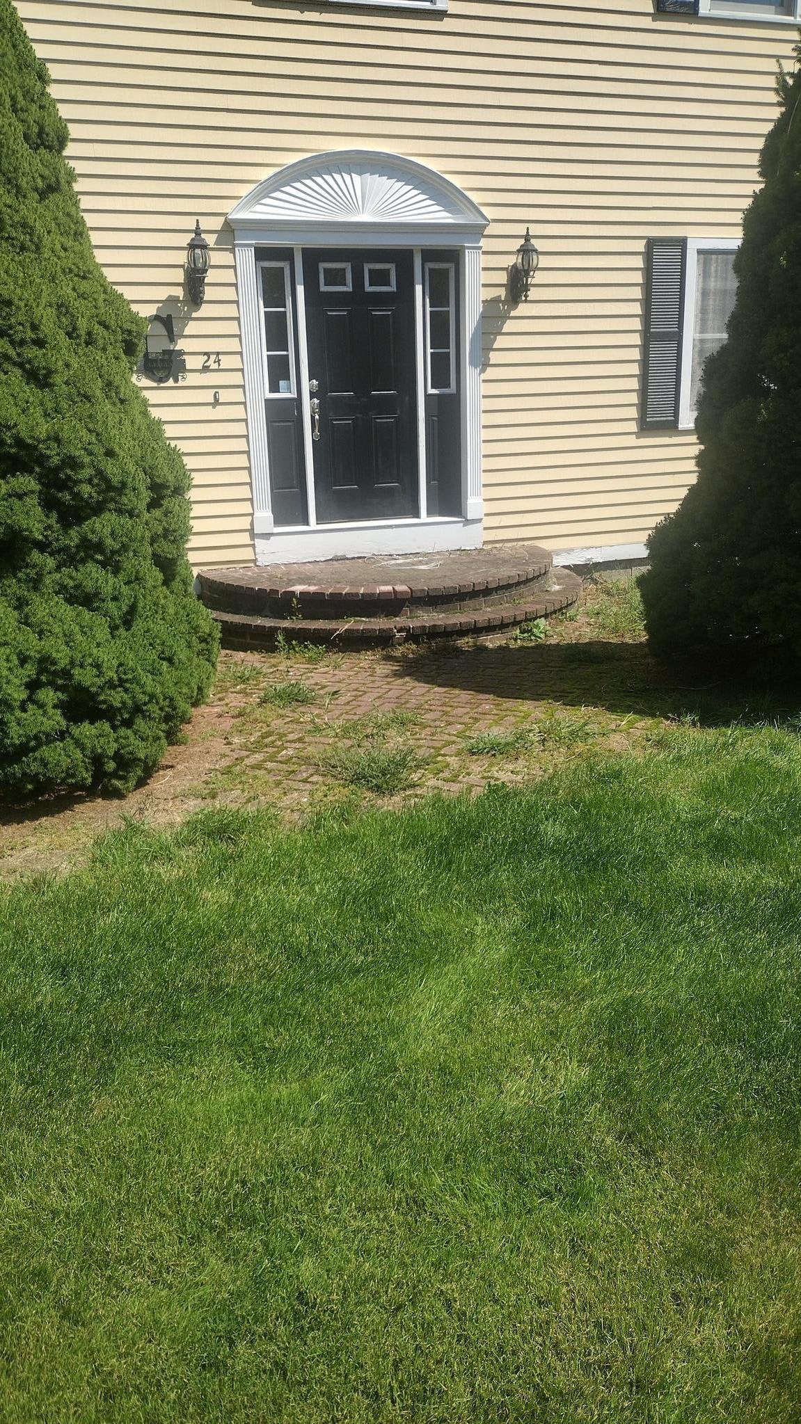 Front entry of a cream-colored house with a black door, semi-circular brick steps, and dense green shrubs, bordered by a weathered brick walkway and a vibrant lawn in Charlton, MA.