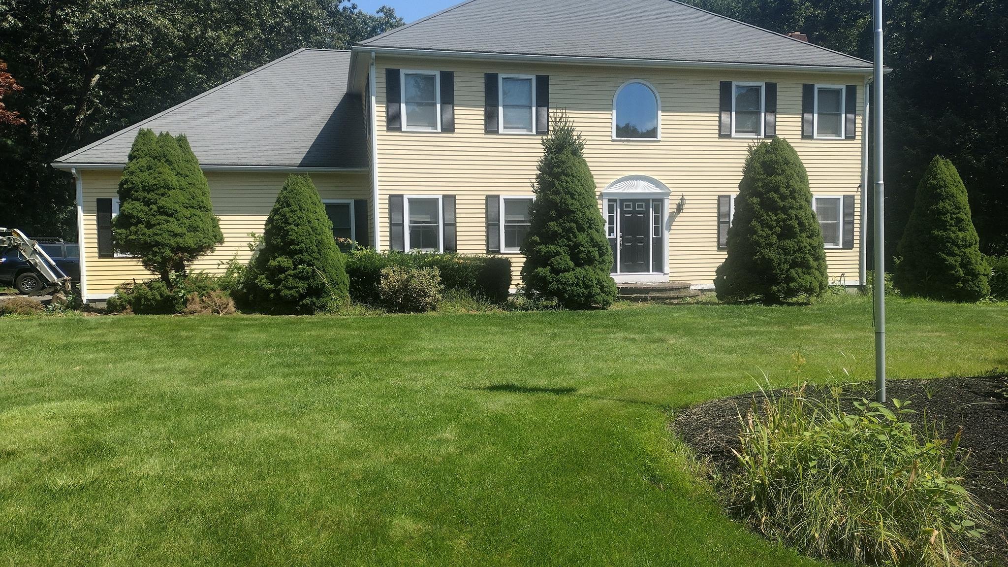 Colonial-style two-story house with light yellow siding, dark shutters, and a well-maintained lawn, surrounded by symmetrical evergreen trees and a flagpole on a sunny day in Charlton, MA.
