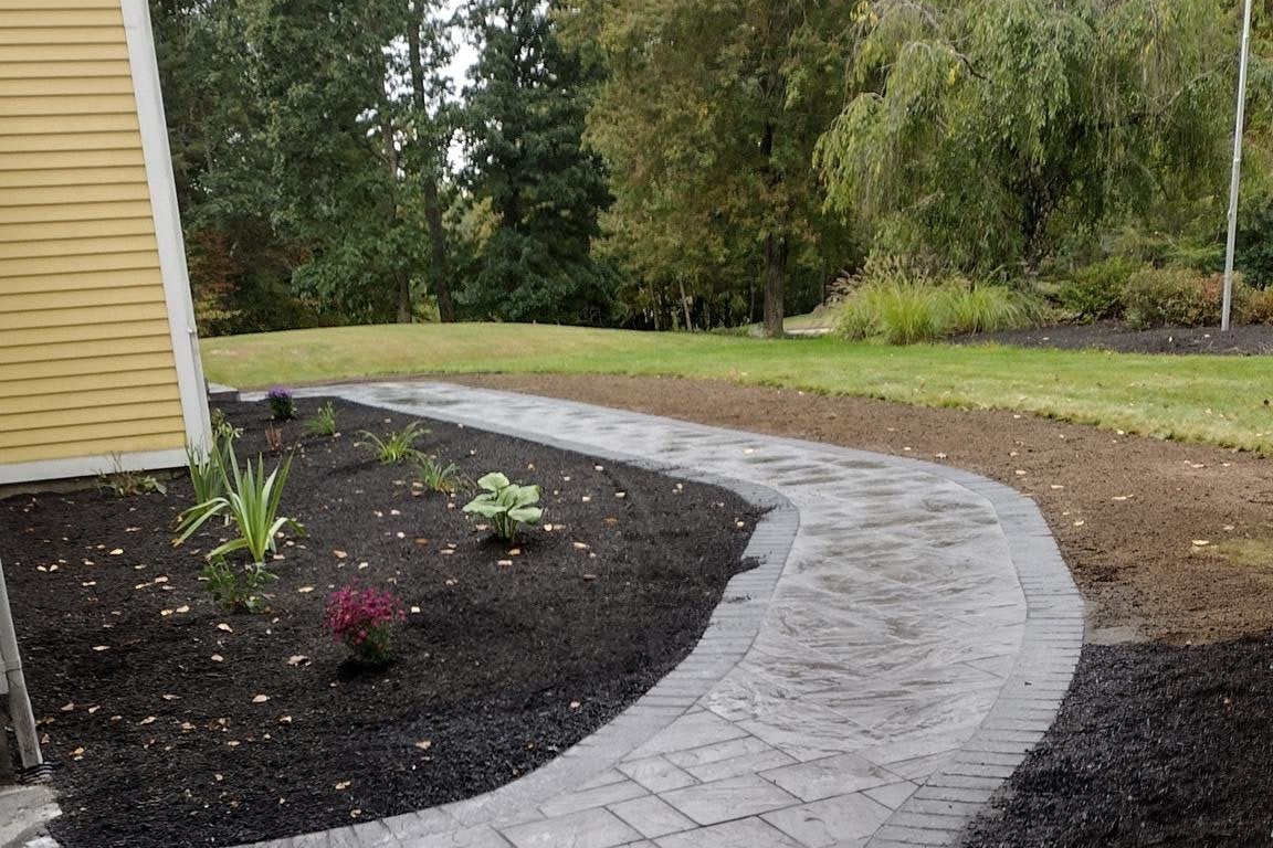 S-curved stamped concrete walkway connecting a dark asphalt driveway to a yellow-sided house in Charlton, MA, surrounded by fresh black mulch, small plants, green grass, and a backdrop of dense trees.