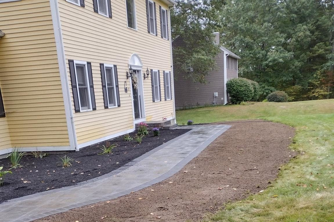 Residential landscaping project in Charlton, MA showing a yellow house with a newly constructed dark gray paver walkway, tilled soil, and survey flags indicating ongoing work.