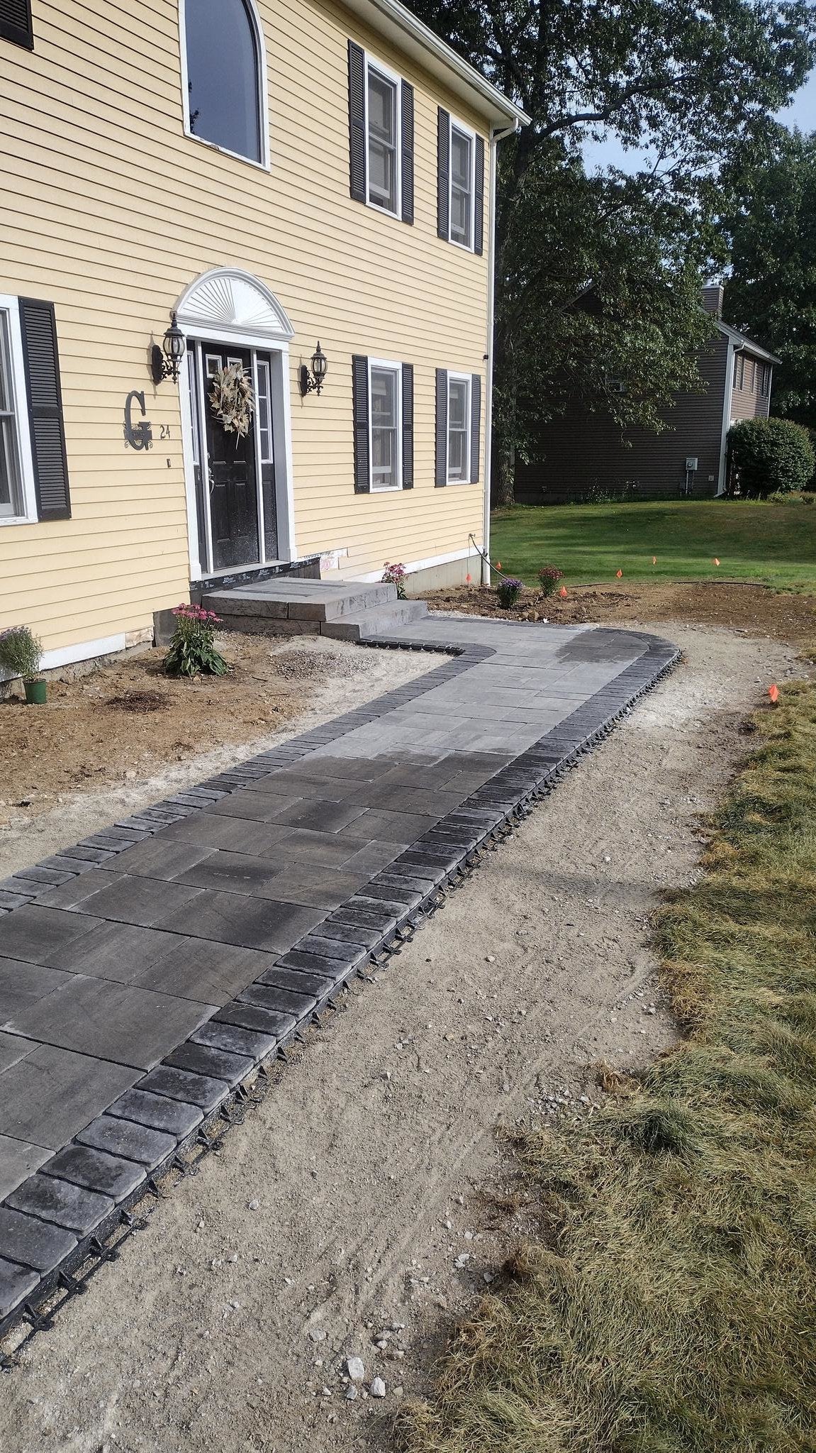 Residential yard in Charlton, MA with a newly installed curved stone walkway, tilled soil for planting, fresh mulch, and a light yellow house surrounded by mature trees.