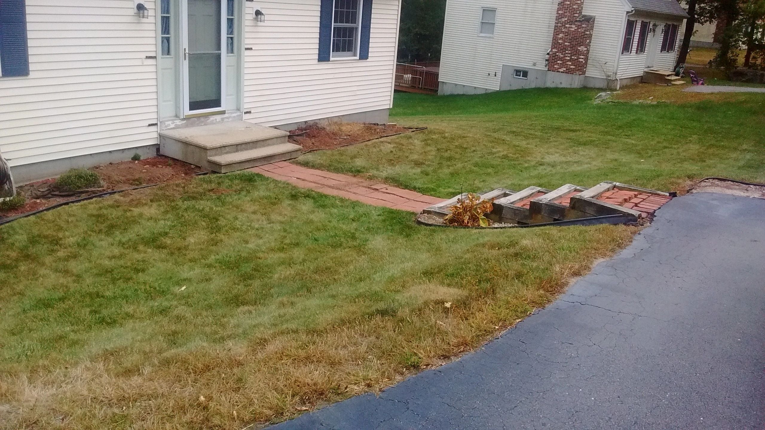 Worn-out concrete steps and a brick walkway in Webster, MA, leading to a house with light siding and blue shutters, surrounded by patchy grass and mulch beds. Project by KRD Landscaping.