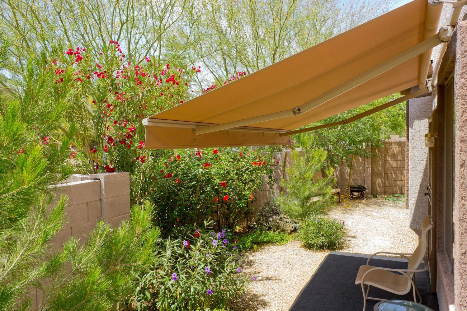 Modern backyard patio with retractable tan awning, ornamental grasses, red dianthus flowers, and purple flowering plants against brick walls on a gravel courtyard.